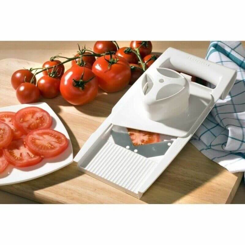 Planche à découper avec trancheuse à tomates et tomates fraîches sur le plan de travail, quelques tranches sur une assiette.