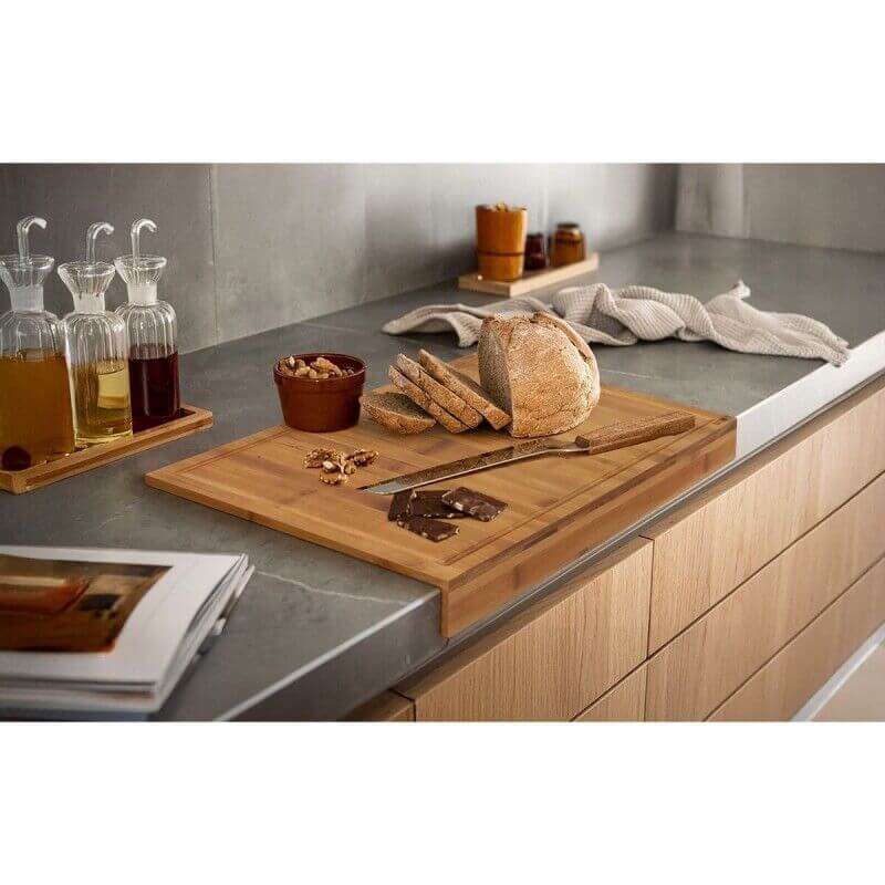 Plan de travail en cuisine avec pain, morceaux de chocolat, couteau et planche à découper en bois, flacons d’huiles et épices en arrière-plan.