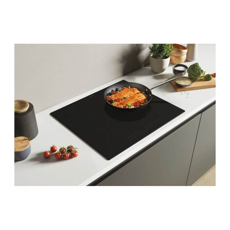 Plaque de cuisson à induction noire avec poêle en cuisson, légumes et tomates cerises à côté, sur plan de travail blanc élégant.