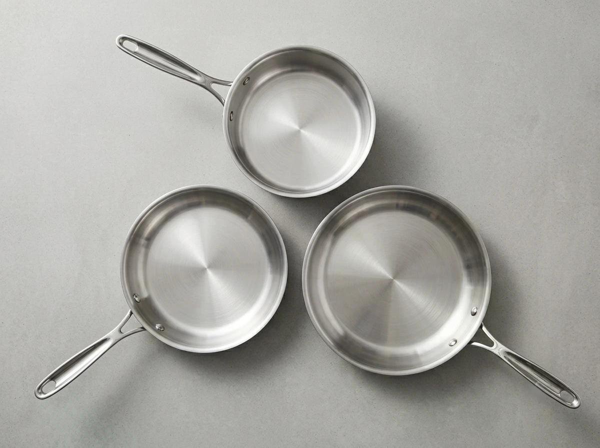 Ensemble de trois petites casseroles en acier inoxydable avec poignées, disposées en triangle sur fond gris.