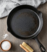 Poêle en fonte noire légèrement rincée avec traces de cuisson, crayon et sel en coin et brosse à vaisselle à côté sur plan de travail en bois.