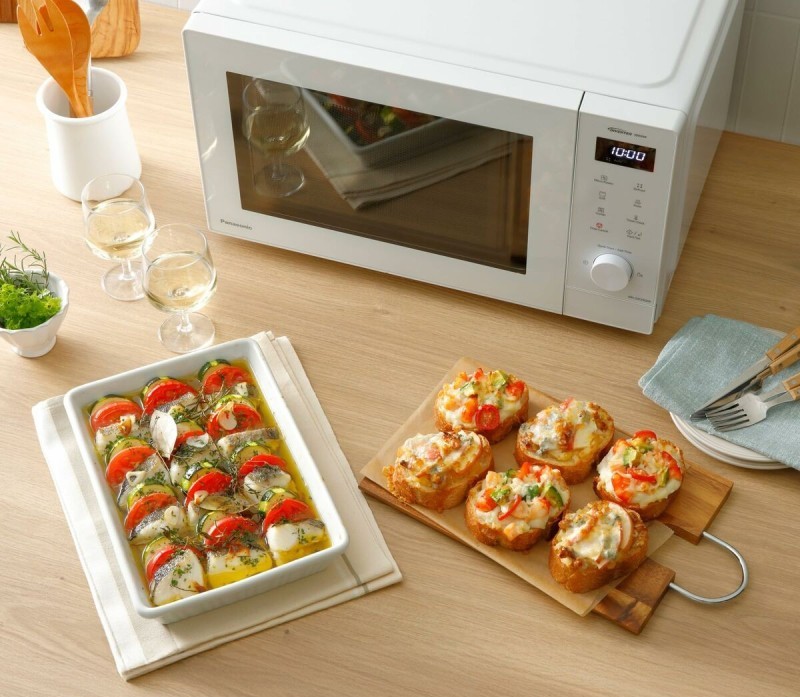 Mikro-ondes blanc sur plan de travail avec plats près: tomates et fromage gratinés dans un plat, canapés/mini-tartines sur planche, verres de vin et ustensiles.